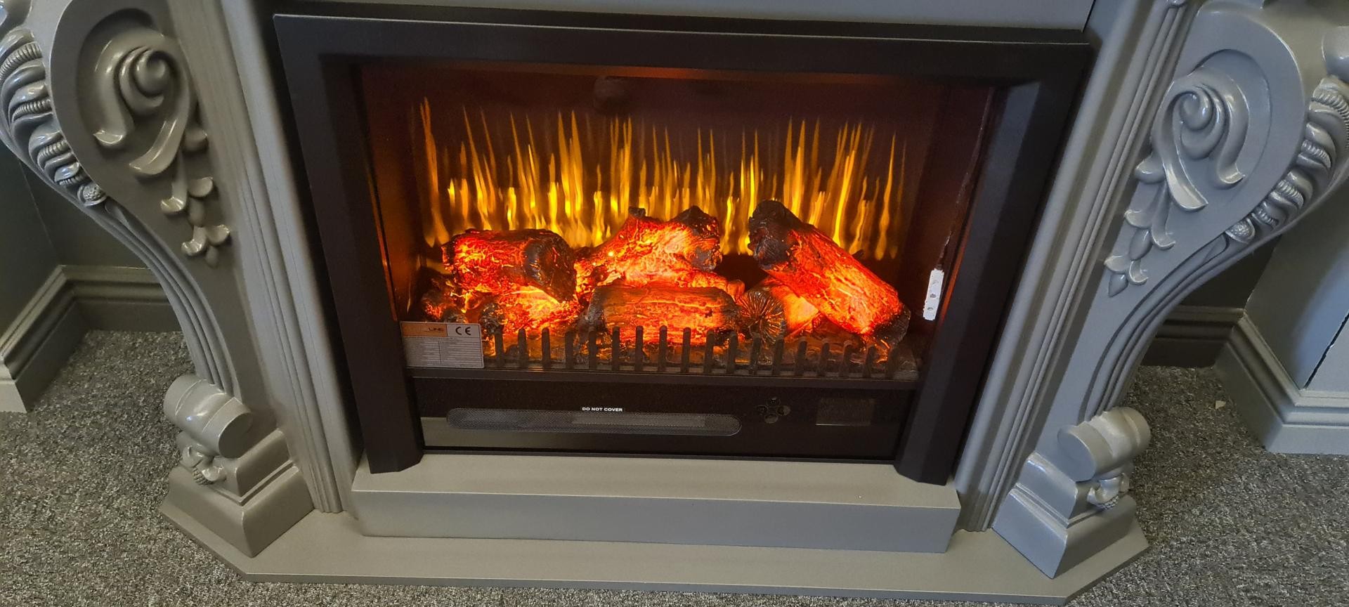 Close-up of a satin grey polyurethane fireplace surround with carved scroll columns framing a glowing electric flame log insert.