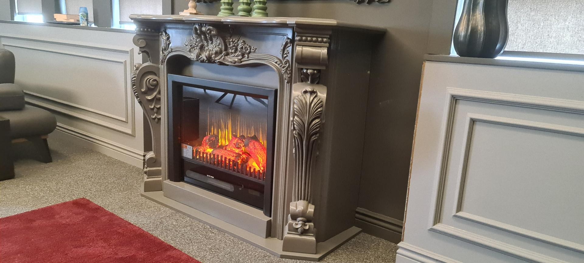 Side angle view of a grey polyurethane fireplace mantle showing deep carved acanthus corbels and electric fireplace glow.