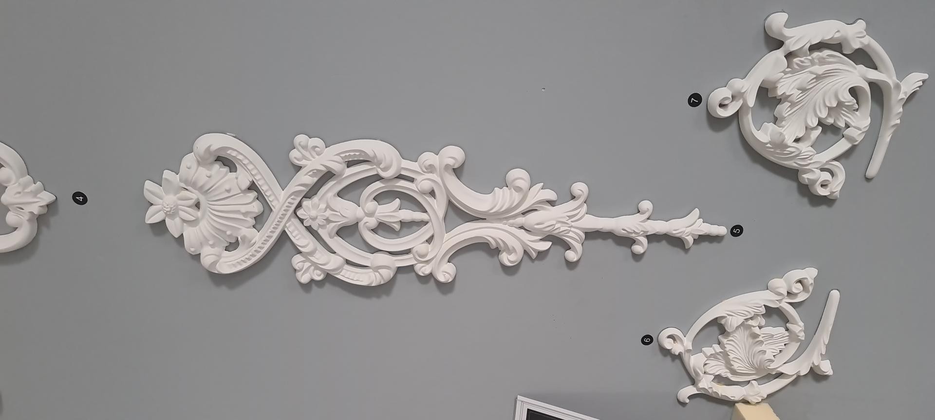 White carved polyurethane wall appliqués including vertical floral drop and corner scroll motifs on a grey wall.