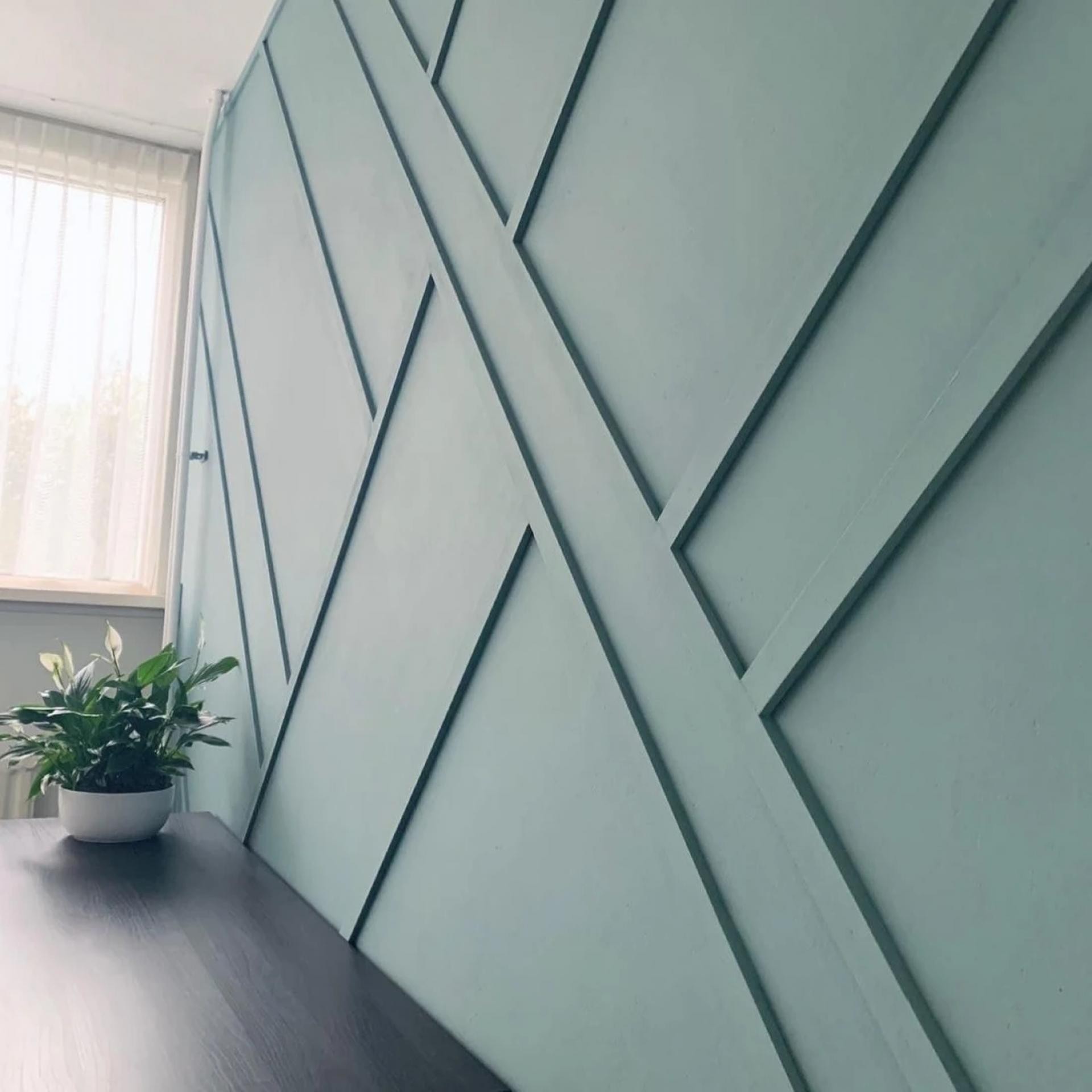 Matte sage green geometric wall paneling created with intersecting diagonal moulding strips beside a dark wood desk.