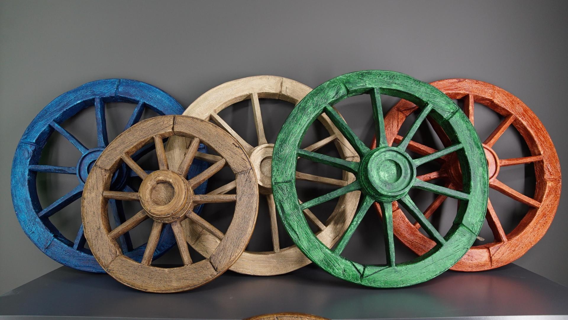 Five overlapping polyurethane wagon wheels painted in blue, beige, green, orange, and natural wood brown on a shelf.