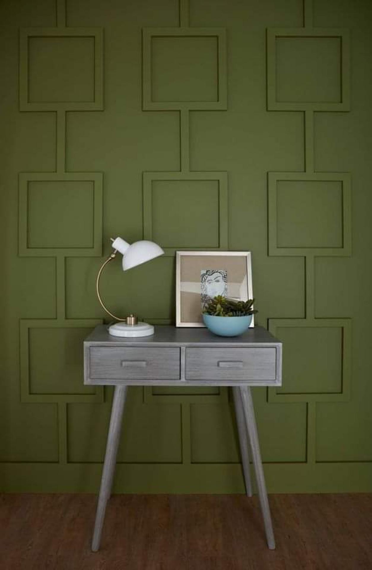 Olive green wall with a geometric square molding pattern, a grey wooden desk, a white task lamp, and a framed abstract art print.