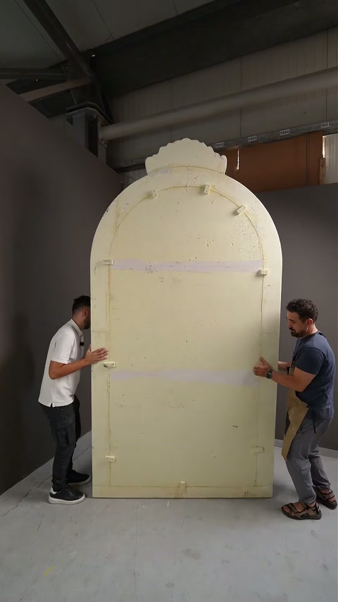 Dois artesãos carregam o suporte de arco inacabado através do espaço de produção da oficina.