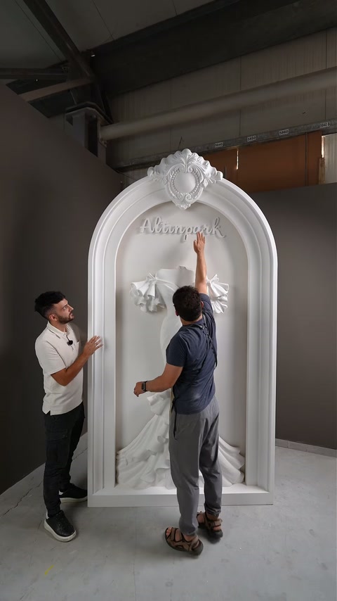 Dois artesãos examinam o posicionamento das letras acima da escultura central de vestido de noiva.