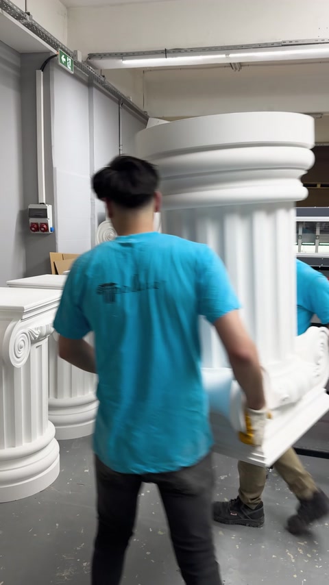 Pracovníci přenášejí velký dekorativní sloup výrobním provozem.