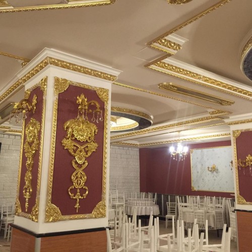 Klassisches Hochzeitssaal-Design mit Polyurethan-Ornamenten und goldenen Details <h2>Klassische Eleganz mit Polyurethan und Gold</h2><p>Der Hochzeitssaal bietet eine prachtvolle Gestaltung, die durch reich verzierte <strong>Polyurethan</strong>-Verkleidungen an Säulen und Decken betont wird. Diese Dekorelemente sind in leuchtendem <strong>Gold</strong> gehalten und kontrastieren elegant mit tiefroten Wandpaneelen. Besonders hervorzuheben sind die floralen Reliefmotive, die wie barocke Verzierungen wirken und dem Raum eine besondere Authentizität verleihen.</p><p>Die Decke ist mit geometrischen Rahmen und symmetrisch angeordneten Ornamentleisten ausgestattet, die den Blick lenken und durch ihre detailreiche Gestaltung für ein harmonisches Gesamtbild sorgen. Ergänzt werden diese Elemente durch üppige Kronleuchter, deren Licht sich auf glänzenden Oberflächen spiegelt und das Ambiente veredelt.</p><h2>Atmosphäre und Funktion</h2><p>Runde Tische und weiße Stühle sorgen für eine klare, elegante Sitzordnung, die in Balance mit der dekorativen Ausdrucksstärke der Architektur steht. Der Tanzbereich bleibt durch die gezielte Anordnung der Möbel frei und schafft ein Zentrum für Bewegung und feierliche Momente.</p><p>Historisch lassen sich solche Ornamente auf barocke Innenarchitekturtraditionen zurückführen, in denen reiche Stuckarbeiten und vergoldete Polyurethanformen – damals in Gips oder Holz umgesetzt – Symbole des Wohlstands und der Feierkultur waren. Diese Neuinterpretation in modernen Materialien verbindet Nostalgie mit praktischer Anwendbarkeit.</p><ul><li>Symmetrische Ornamentleisten</li><li>Goldene florale Reliefs</li><li>Polyurethan-Profile mit hoher Detailgenauigkeit</li><li>Kombination aus Rot, Weiß und Gold</li></ul>
<p>Dieser Hochzeitssaal zeigt eine opulente Gestaltung, die klassische Formen neu interpretiert. Die Verwendung von <strong>Polyurethan</strong>-Verzierungen sorgt für raffinierte Details. Goldene Ornamente auf rotem Hintergrund betonen die luxuriöse Wirkung. Weiße Stühle und Tische bilden einen eleganten Kontrast. Symmetrische Linien verstärken die klare Raumwirkung. Kronleuchter bringen eine warme Atmosphäre. Wandappliken ergänzen das Beleuchtungskonzept. Florale <strong>Reliefs</strong> steigern die dekorative Tiefe. Profilierte Deckenleisten schaffen Struktur und Harmonie. Der freie Tanzbereich bleibt im Zentrum des Designs. Die Farbpalette aus Rot, Weiß und <strong>Gold</strong> erzeugt feierliche Stimmung. Historische Einflüsse barocker Innenräume sind erkennbar. Moderne Materialien machen die Gestaltung langlebig. Gäste erleben eine Balance aus Pracht und Funktionalität. Insgesamt entsteht ein klassisches Ambiente für unvergessliche Feste.</p>