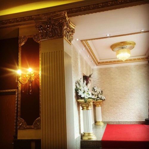 Klassischer Hochzeitssaal mit Polyurethan-Säulen und vergoldeten Ornamenten <h2>Klassische Säulenarchitektur</h2><p>Der Hochzeitssaal präsentiert sich in einem prunkvollen Stil, in dem <strong>Polyurethan</strong>-Säulen mit vergoldeten Kapitellen dominieren. Diese sind reich verziert, inspiriert von <strong>Korinthischen</strong> Details, die historisch schon in antiken römischen Bauten verwendet wurden. Die vertikalen Rillen der Säulen erzeugen eine harmonische Balance und betonen die <strong>Symmetrie</strong> im Innenraum.</p><h2>Dekorative Details und Materialien</h2><p>Die Decke ist mit goldfarbenen Stuckleisten eingefasst, die eine klare geometrische Rahmung bilden. Ein Kristalllüster mit goldener Umrahmung verleiht dem Raum zusätzlichen Glanz. Wandapplikationen mit floralen Mustern setzen weiche Akzente im Kontrast zu den klar strukturierten Säulen. Diese Kombination aus <strong>Ornamentik</strong>, Textur und Licht unterstützt ein luxuriöses Ambiente.</p><h2>Gestaltungsideen für Innenarchitekten</h2><p>Innenarchitekten können auf die Wirkung von <strong>Geometrie</strong>, Farbkontrasten und feinen Ornamenten setzen, um klassische Räume modern zu interpretieren. Hierbei spielen nicht nur die Wahl der Materialien, sondern auch die Positionierung von Lichtquellen eine wesentliche Rolle. Eine dezente Wandtextur in Steinoptik und reicher Golddekor schaffen eine Hochzeitssaal-Atmosphäre, die gleichermaßen festlich wie zeitlos ist.</p>

<p>Ein klassischer Hochzeitssaal lebt von detailreicher Ornamentik. Säulen mit <strong>Polyurethan</strong>-Verkleidung schaffen eleganten Glanz. Vergoldete Kapitelle lassen die Architektur monumental wirken. Die vertikalen Rillen der Säulen verstärken die Wahrnehmung der Höhe. Ein Kristalllüster bildet den glanzvollen Mittelpunkt der Decke. Goldene Stuckleisten rahmen die Architektur und unterstreichen die Prunkhaftigkeit. Die Wandflächen sind in dezenter Steinoptik gehalten. Florale Dekorarrangements bringen weiche Kontraste in die Symmetrie. Das kräftige Rot des Teppichs setzt einen festlichen Akzent. Das Zusammenspiel von <strong>Geometrie</strong> und Textur schafft ein harmonisches Gesamtbild. Historische Einflüsse wie <strong>Korinthische</strong> Ornamente verleihen Authentizität. Moderne Materialien wie Polyurethan verbinden Tradition und Beständigkeit. <strong>Ornamentik</strong> und Lichtführung ergeben eine ausgewogene Raumstimmung. Der Hochzeitssaal vereint klassische Eleganz mit zeitloser Festlichkeit. Diese Gestaltung bietet Innenarchitekten vielseitige Anregungen.</p>