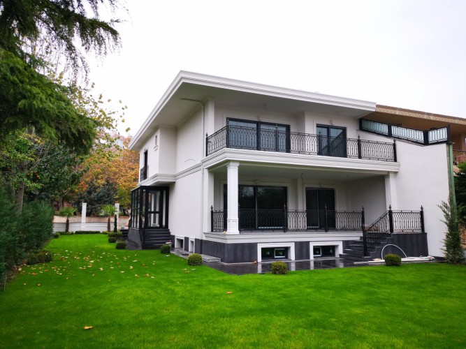 Villa moderne me dekor klasik me poliuretan dhe detaje të fasadës <h2>Dekor klasik me elemente moderne</h2><p>Kjo vilë përdor material <strong>poliuretan</strong> për veshjen e fasadës, duke siguruar rezistencë dhe elegancë njëkohësisht. Shtyllat e bardha mbajnë erën e <strong>stil klasik</strong> dhe kujtojnë elementë të arkitekturës së lashtë, duke sjellë një ndjesi madhështie dhe përmasë të harmonishme.</p><p>Kornizat horizontale dhe basorelievet e thjeshta ndërthuren me forma të drejta, duke e bërë ndërtesën të marrë një karakter <strong>modern</strong>. Balustradat prej hekuri të zi janë zbukuruar me detaje simetrike, duke krijuar ritëm dekorativ me motive të përsëritura lineare dhe gjeometrike.</p><p>Pjesa e poshtme e fasadës me ngjyrë gri të errët krijon një bazament vizual që e shkëput vëllimin e bardhë të ndërtesës, duke shtuar thellësi kompozicionit. Ky detaj është i rëndësishëm në traditën klasike, ku bazamenti kishte një funksion simbolik të forcës dhe qëndrueshmërisë.</p><p>Hapësira e gjelbër me shkurre sferike dhe bar të dendur jeshil plotëson ndërtesën, duke theksuar kontrastin natyrë–arkitekturë. Simetria e fasadës dhe ekuilibri ndërmjet dritareve ndihmojnë në ruajtjen e harmonie vizuale.</p><ul><li>Përdorimi i <strong>poliuretanit</strong> për kornizat dhe dekorimet</li><li>Kombinimi i ngjyrës së bardhë me detaje të zeza dekorative</li><li>Shtyllat vertikale klasike që kujtojnë rendet antike</li><li>Kopsht i gjelbër i rregulluar me forma gjeometrike</li></ul><p>Arkitektura përçon një lidhje mes traditës dhe modernitetit, përmes dritareve të mëdha, shtyllave klasike dhe dekorimit me lirizëm bashkëkohor.</p>

<p>Kjo vilë është një shembull i ndërthurjes së estetikës moderne dhe klasike. Fasada e saj përdor <strong>poliuretan</strong> për qëndrueshmëri dhe finesë. Ngjyra e bardhë e mureve thekson pastërtinë vizuale. Shtyllat vertikale sjellin ndjenjën e monumentalitetit. Kornizat horizontale krijojnë një rrafsh elegant dhe të balancuar. Rethojat metalike të zeza shtojnë elementin grafik. Motivet gjeometrike të tyre janë ritmike dhe të sakta. Dyshemeja e errët nxjerr në pah bazamentin e vilës. Shkurret sferike japin një ndjesi disipline vizuale. Kombinimi i bari të gjelbër dhe fasadës së bardhë gjallëron pamjen. Dritaret e mëdha sjellin dritë natyrale brenda. Ndërtesa përfaqëson elegancën funksionale. Kjo qasje arkitekturore i jep jetë një harmoni estetike. Vila ilustron një ekuilibër të veçantë mes traditës dhe inovacionit. Ky dizajn është frymëzim për dekorime të ardhshme.</p>