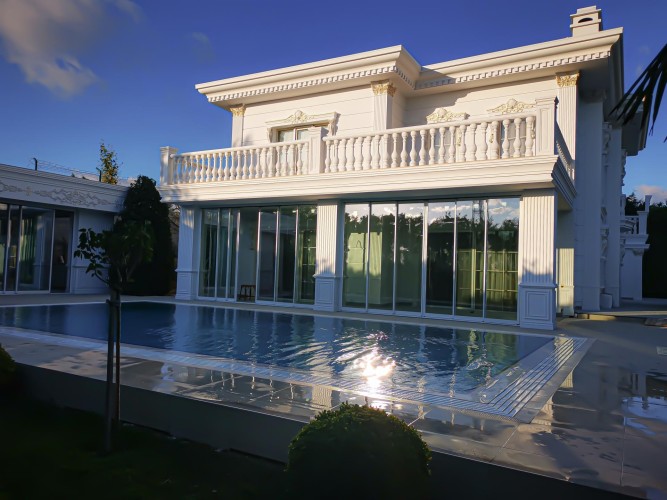 Villa classica con dettagli in poliuretano per una facciata elegante e durevole <h2>Eleganza e resistenza nel design classico</h2><p>Il poliuretano applicato alle facciate permette di creare decorazioni raffinate che ricordano materiali antichi, ma con maggiore praticità. Le <strong>colonne</strong> scanalate, i fregi e le balaustre in poliuretano offrono un risultato estetico di grande qualità, mantenendo leggerezza strutturale e resistenza nel tempo.</p><p>L’uso di <strong>poliuretano</strong> nelle rifiniture esterne offre la possibilità di inserire motivi ornamentali tradizionali senza gravare sulle strutture portanti. Questo materiale resiste all’umidità, al sole e alle variazioni climatiche, preservando l’armonia visiva della villa.</p><h2>Dettagli decorativi e motivi geometrici</h2><p>La facciata mostra un equilibrio tra rigore architettonico e dettagli ornatissimi. Le cornici orizzontali sottolineano la geometria, mentre i fregi dorati aggiungono un tocco di prestigio e raffinatezza. Le <strong>balaustre</strong> proporzionate creano un ritmo armonioso lungo il balcone.</p><p>Gli ornamenti richiamano elementi della tradizione architettonica classica: la verticalità delle colonne ricorda i modelli ionici e corinzi che per secoli hanno ispirato la costruzione di ville e palazzi. Questo richiamo storico conferisce alla villa un carattere senza tempo.</p><h2>Un dialogo con l’ambiente</h2><p>La piscina riflette i motivi lineari della struttura, creando un continuum visivo. Gli spazi in vetro trasparente favoriscono la luce naturale e rendono gli interni in connessione con l’esterno. L’uso del <strong>bianco</strong> come tonalità dominante, con dettagli dorati, amplifica l’effetto di luminosità e maestosità.</p><ul><li>Motivi geometrici evidenziati con cornici e fregi</li><li>Balaustre tornite e simmetriche</li><li>Colonne con scanalature verticali</li><li>Dettagli dorati che valorizzano gli angoli</li></ul><p>Grazie a questi elementi, la villa assume un valore estetico che unisce <strong>tradizione</strong> e innovazione tecnologica.</p>
<p>Questa villa con decorazioni in poliuretano unisce eleganza e durata. Le colonne scanalate ricordano l’architettura classica. Le balaustre creano ritmo e armonia visiva. I fregi dorati aggiungono prestigio senza eccesso. Il bianco dominante amplifica la luminosità. La piscina integra lo spazio con riflessi geometrici. Le superfici lucide donano profondità. Il poliuretano garantisce leggerezza e resistenza. I dettagli proporzionati esaltano la simmetria. La tradizione classica si coniuga con materiali moderni. Le cornici orizzontali rafforzano la geometria. Ogni ornamento sottolinea la qualità artigianale. L’uso del <strong>poliuretano</strong> preserva estetica e funzionalità. Le <strong>colonne</strong> e le <strong>balaustre</strong> raffinate trasmettono solidità. Questa combinazione offre una villa iconica e senza tempo.</p>