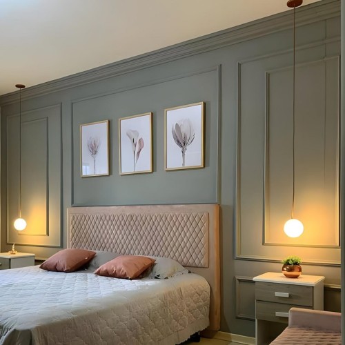 Minimalistische Wandvertäfelung mit Polyurethan im modernen klassischen Schlafzimmer <h2>Eleganz im modernen klassischen Schlafzimmer</h2><p>Die Verwendung von <strong>Polyurethan</strong>-Paneelen für die Wandvertäfelung schafft klare Linien und eine subtile Struktur, die den Raum sowohl modern als auch klassisch wirken lässt. Die rechteckigen Einfassungen erinnern an historische Wandverkleidungen, wie sie bereits in europäischen Herrenhäusern verwendet wurden.</p><p>Ein weicher Kopfteil mit <strong>rautenförmigem</strong> Steppmuster harmoniert perfekt mit den streng geometrischen Wandrahmen und bringt Wärme und Komfort ins Design. Die harmonische Farbpalette in <strong>Salbeigrün</strong> und Beige lässt den Raum ruhig und ausgewogen erscheinen.</p><p>Die drei floralen Bilder in goldenen Rahmen über dem Bett setzen einen leichten künstlerischen Akzent und ergänzen die symmetrische Anordnung. Ebenso tragen die schwebenden Nachttische mit schlichten Linien zur <strong>minimalistischen</strong> Wirkung bei.</p><p>Besondere Aufmerksamkeit verdienen die hängenden Kugelleuchten, die mit ihrem gleichmäßigen Licht eine <strong>atmosphärische</strong> Stimmung erzeugen. Durch ihre vertikale Ausrichtung unterstreichen sie zusätzlich das Spiel aus Höhe und Geometrie.</p><ul><li>Symmetrische Wandvertäfelungen als Raumstruktur</li><li>Geometrisches Steppmuster am Kopfteil</li><li>Zarte florale Drucke im Goldrahmen</li><li>Kugelförmige Hängelampen mit warmem Licht</li></ul><p>Die Gestaltung zeigt, wie klassisches Dekor durch neue Materialien wie Polyurethan in moderne Innenräume integriert werden kann. Diese Kombination führt zu einem zeitlosen Interieur mit starker ästhetischer Ausstrahlung.</p>
<p>Dieses Schlafzimmerdesign kombiniert klassische Wandvertäfelung mit moderner Zurückhaltung. Die <strong>Polyurethan</strong>-Paneele erzeugen klare Geometrien. Der rautenförmige Kopfteil betont Struktur und Komfort. Zarte Grün- und Beigetöne schaffen Ruhe und Balance. Goldgerahmte florale Bilder fügen dezente Eleganz hinzu. Historische Anklänge finden eine zeitgenössische Interpretation. Die <strong>minimalistische</strong> Gestaltung strahlt Harmonie aus. Pendelleuchten mit Kugelform setzen warme Lichtpunkte. Symmetrie bestimmt den gesamten Aufbau. Geometrische Details verstärken die vertikale Wirkung. Fein abgestimmte Farbtöne wirken beruhigend. Durch die Kombination von Klassik und Moderne entsteht ein zeitloser Raum. Der Einsatz von <strong>atmosphärischem</strong> Licht steigert die Behaglichkeit. Die Blumenmotive bringen Leichtigkeit. Insgesamt überzeugt das Design durch klare Linien, <strong>Eleganz</strong> und nachhaltige Raffinesse.</p>