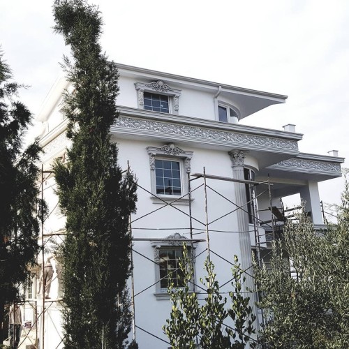 Villa clássica com fachada em poliuretano e detalhes elegantes <h2>Design Clássico com Poliuretano</h2><p>O uso de <strong>poliuretano</strong> na fachada desta villa garante resistência às intempéries e leveza nas formas ornamentais. As molduras envolventes das janelas exibem <strong>detalhes esculpidos</strong> que remetem a arabescos tradicionais, criando um contraste elegante com a lisura das paredes brancas.</p><p>As <strong>colunas clássicas</strong> reforçam a imponência da estrutura, trazendo inspiração da tradição grega e romana, especialmente observada no formato dos capitéis com linhas delicadas e ornamentos em relevo. Esse recurso arquitetônico, muito utilizado desde a Antiguidade, ainda hoje transmite prestígio e sofisticação.</p><h2>Detalhes Ornamentais e Estética</h2><p>A cornija que percorre horizontalmente o perímetro da casa destaca-se pelo conjunto de <strong>motivos decorativos</strong> simétricos, em estilo floral-geometrizado, proporcionando unidade visual. Já as molduras retangulares das janelas são suavizadas por arabescos superiores que quebram a rigidez das linhas.</p><ul><li>Molduras com arabescos e curvas refinadas</li><li>Perfis de poliuretano em alto relevo</li><li>Colunas cilíndricas com capitéis detalhados</li><li>Cornija contínua com decoração clássica</li></ul><p>A paleta cromática em branco e cinza realça os ornamentos, oferecendo um aspecto sofisticado e atemporal.</p>

<p>Esta villa exibe uma fachada em <strong>poliuretano</strong> que combina resistência e elegância. Os ornamentos ao redor das janelas mostram <strong>detalhes esculpidos</strong> inspirados em arabescos clássicos. As <strong>colunas clássicas</strong> reforçam a imponência da arquitetura. A cornija contínua apresenta riqueza de <strong>motivos decorativos</strong> com precisão. O contraste entre paredes lisas e relevos ornamentais cria sofisticação. As molduras envolvem as aberturas com delicadeza floral. O acabamento em branco com cinza adiciona leveza ao conjunto. A volumetria é realçada pelas linhas retas em equilíbrio com curvas. Os elementos em relevo permitem um jogo de luz e sombra refinado. A inspiração greco-romana aparece em cada detalhe estrutural. A simetria dos ornamentos garante harmonia visual. O design une tradição e modernidade em equilíbrio. Os relevos geométrico-florais acrescentam textura estética. A escolha do poliuretano assegura durabilidade e leveza. O resultado é uma composição elegante com caráter atemporal.</p>