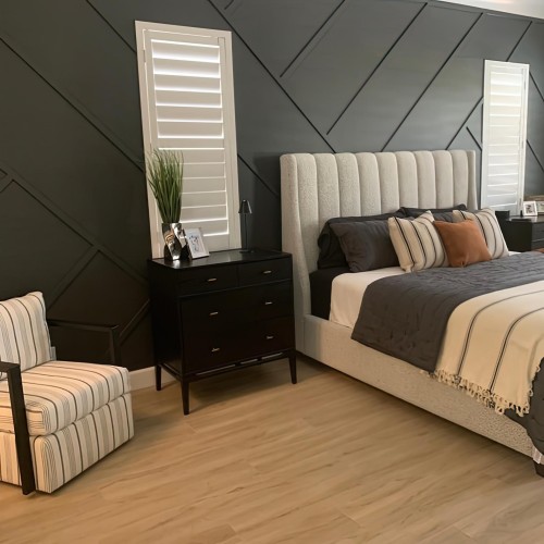 Camera moderna con parete geometrica scura e dettagli classici <h2>Parete geometrica e modanature in poliuretano</h2><p>La parete principale della stanza è arricchita da <strong>modanature</strong> in rilievo disposte in diagonale, realizzate in <strong>poliuretano</strong> resistente, che donano profondità e ritmo visivo. Questo motivo geometrico non solo decora, ma definisce lo stile complessivo dell’ambiente, combinando modernità e richiami classici.</p><p>Le <strong>persiane</strong> bianche incorniciano con precisione lo schema geometrico scuro, portando luminescenza e contrasto alla stanza. Il disegno lineare e verticale delle persiane genera un equilibrio simmetrico che dialoga con il dinamismo delle linee diagonali.</p><h2>Arredo e materiali</h2><p>Il letto imbottito, con una testata alta rivestita in tessuto chiaro, contrasta elegantemente con il fondo scuro. Sulla superficie del letto, i cuscini con strisce verticali e uno in tinta ramata offrono una varietà cromatica equilibrata. I comodini neri con linee semplici e i piccoli dettagli metallici rafforzano l’atmosfera moderna.</p><p>Il pavimento in <strong>legno</strong> chiaro valorizza l’arredo, introducendo calore e naturalezza. Una poltrona a righe, con tonalità neutre, aggiunge comfort e un tocco accogliente, mantenendo una coerenza con lo schema grafico generale.</p><h2>Un tocco classico nella modernità</h2><p>L’uso delle modanature ricorda brevemente la tradizione architettonica classica, dove gli intarsi e i rilievi davano ritmo agli spazi interni. Qui, tali elementi vengono reinterpretati in chiave moderna, diventando parte integrale dell’estetica contemporanea.</p><ul><li>Linee diagonali che creano movimento.</li><li>Contrasto tra scuro e chiaro per armonia visiva.</li><li>Elementi tessili che ammorbidiscono lo spazio.</li></ul>

<p>Questa camera mostra un raffinato equilibrio tra stile moderno e dettagli classici. Le modanature in <strong>poliuretano</strong> disposte in diagonale sulla parete scura donano movimento. Le <strong>persiane</strong> bianche amplificano la luminosità naturale. L’alto letto imbottito crea un punto focale lussuoso. I cuscini multicolore aggiungono vivacità equilibrata. Il pavimento in <strong>legno</strong> chiaro scalda l’atmosfera. Una poltrona a righe sottolinea la coerenza grafica. I comodini neri rafforzano il contrasto cromatico. La disposizione geometrica garantisce rigore architettonico. L’unione di ombre e luci definisce tridimensionalità. Grazie alla scelta dei materiali, la stanza appare armoniosa. I tessuti morbidi bilanciano la rigidità delle linee. La composizione complessiva unisce funzionalità ed estetica. Richiami classici aggiungono valore storico alla modernità. La combinazione di colori neutri garantisce eleganza duratura. Ogni dettaglio contribuisce a un design sofisticato e rilassante.</p>